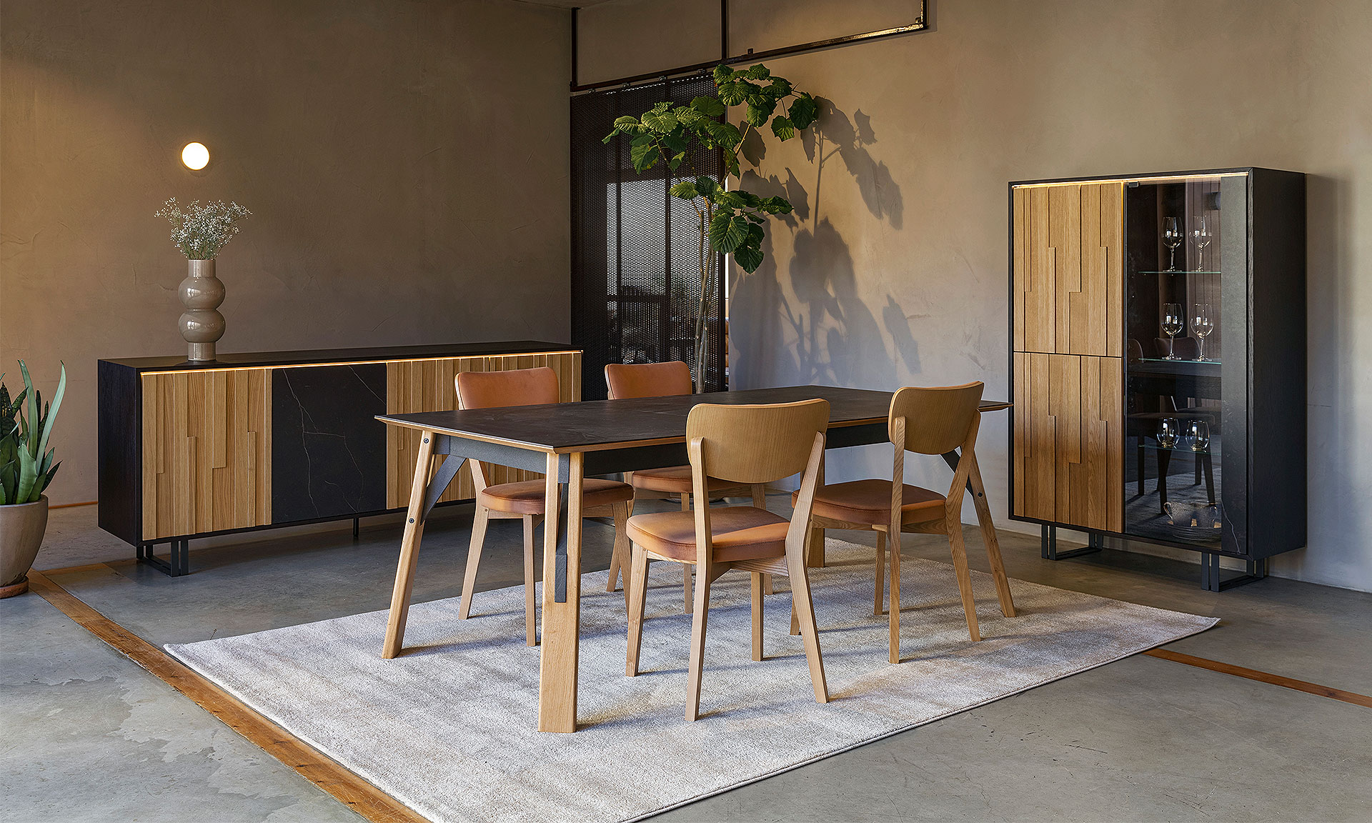 Photo d'une salle à manger avec une collection comportant un buffet, une table, une vitrine haute et des chaises. Le meuble présente des finitions en métal noir mat et chêne en relief.